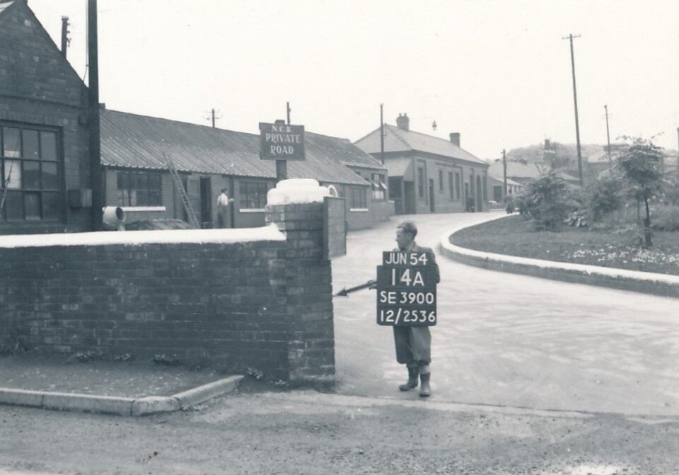 A black-and-white photo shows a young person holding a sign with survey details near a brick wall on a quiet, curving street lined with industrial buildings and houses. The sign reads “N.C.B Private Road.”.